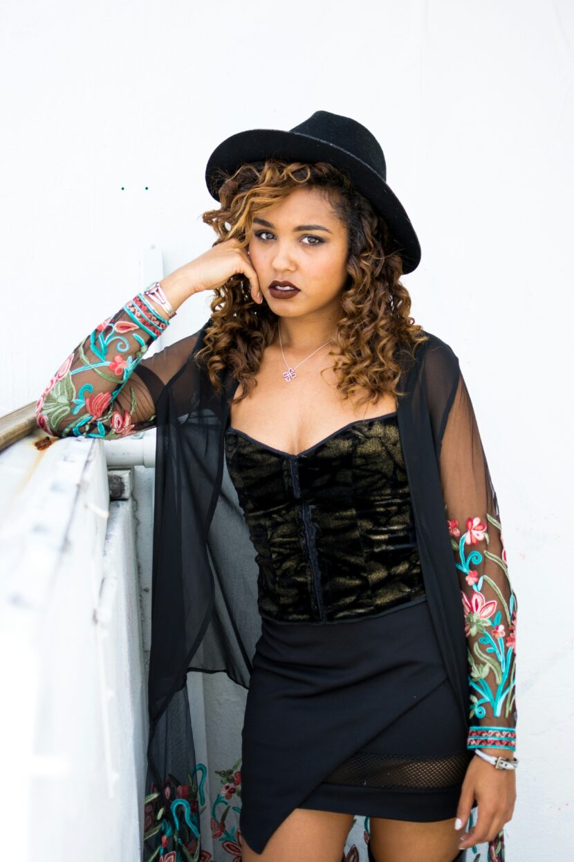 Miami Fashion: The Ten Iconic Items To Rock This Summer Young woman with curly hair leans on a white railing. She's wearing a black hat, sheer embroidered black shawl, and dark lipstick, exuding a confident, stylish vibe.