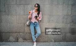 Styling Your Pants For Fashionable Girls A stylish woman in sunglasses leans against a textured stone wall, wearing a pink jacket, blue jeans, and white sneakers, holding a drink with a smile.