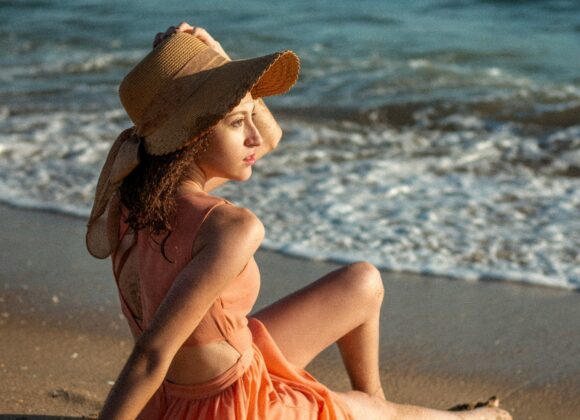 The 10 Best Beach Dresses For The Summer Woman in a peach dress and straw hat sits on a sunlit beach, gazing at the ocean waves. The scene conveys a peaceful and relaxed summer mood.