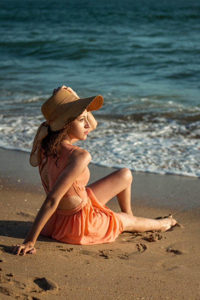 The 10 Best Beach Dresses For The Summer Woman in a peach dress and straw hat sits on a sunlit beach, gazing at the ocean waves. The scene conveys a peaceful and relaxed summer mood.