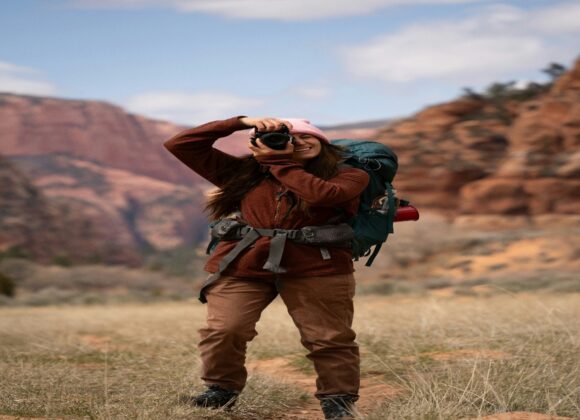 🏜️ Grand Canyon Clothing Tips: What to Wear for Style & Survival A woman in hiking gear stands smiling, taking a photo with a camera. She's in a scenic canyon with red rock formations and a bright, cloudy sky.