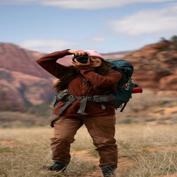 A woman in hiking gear stands smiling, taking a photo with a camera. She's in a scenic canyon with red rock formations and a bright, cloudy sky.