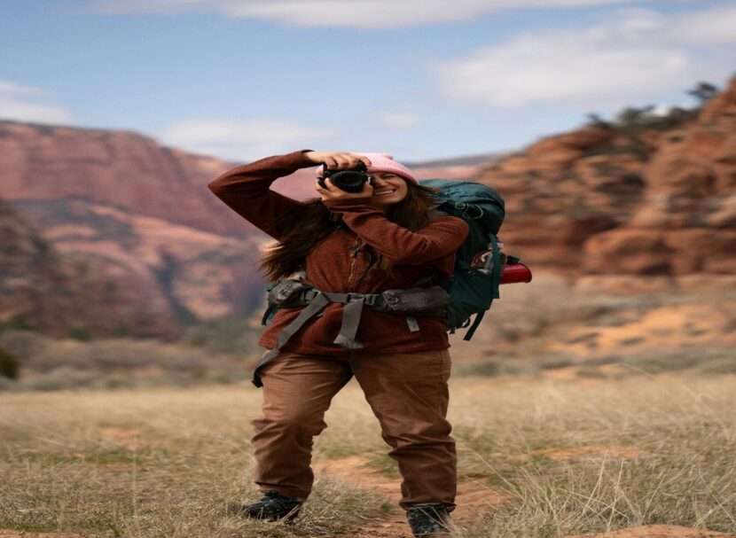 A woman in hiking gear stands smiling, taking a photo with a camera. She's in a scenic canyon with red rock formations and a bright, cloudy sky.