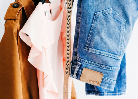 Best Denim Jacket for Women (2025): Why a Super Soft Jean Jacket Belongs in Every Closet A light brown jacket, a pink ruffled top, and blue jeans hang on a rack. A beige belt with a pattern is draped over the rail, suggesting a casual style.