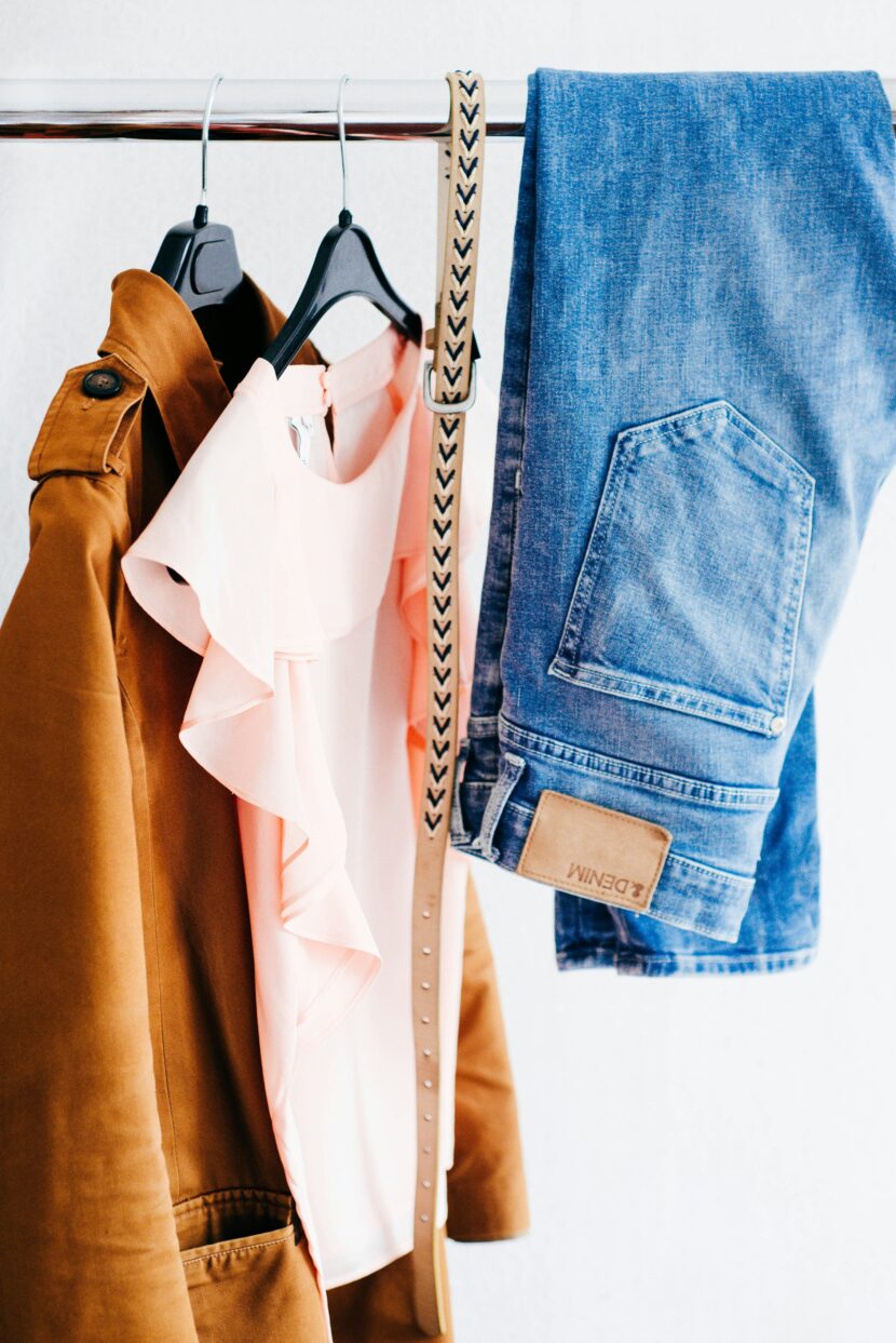 Best Denim Jacket for Women (2025): Why a Super Soft Jean Jacket Belongs in Every Closet A light brown jacket, a pink ruffled top, and blue jeans hang on a rack. A beige belt with a pattern is draped over the rail, suggesting a casual style.