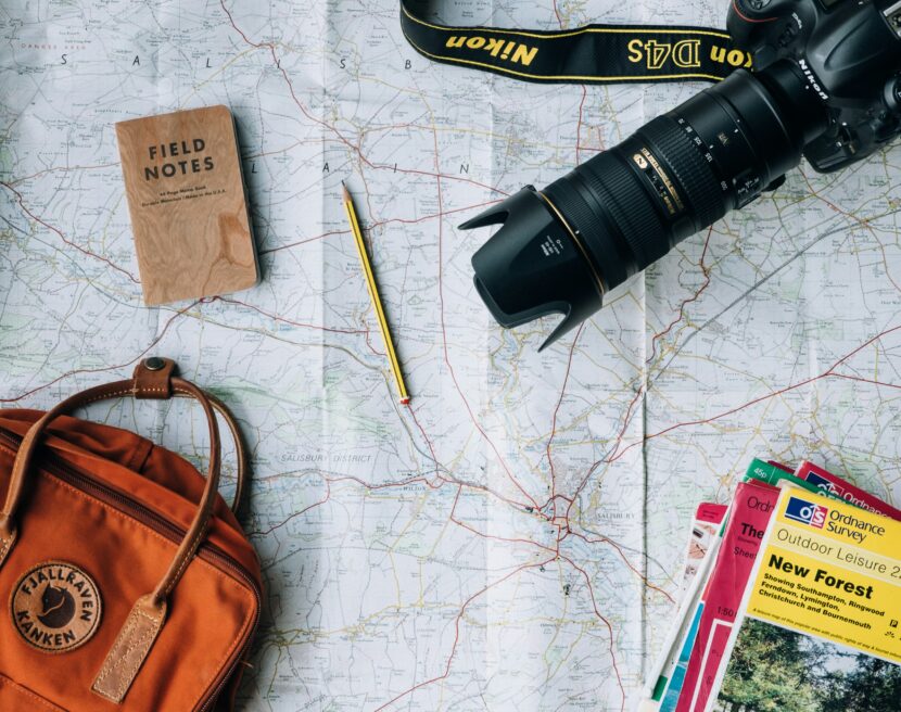 A travel-themed flat lay featuring a map, camera, notebook, pencil, orange backpack, and colorful guidebooks, evoking a sense of adventure.