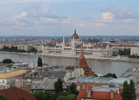 What to Wear in Budapest in September View of the Hungarian Parliament Building alongside the Danube River under a partly cloudy sky. Foreground shows city rooftops, evoking a serene urban landscape.