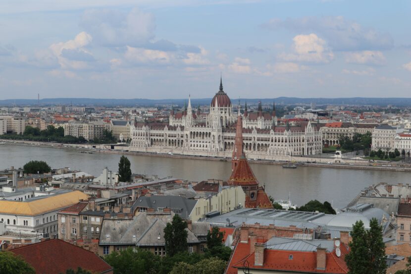 What to Wear in Budapest in September View of the Hungarian Parliament Building alongside the Danube River under a partly cloudy sky. Foreground shows city rooftops, evoking a serene urban landscape.