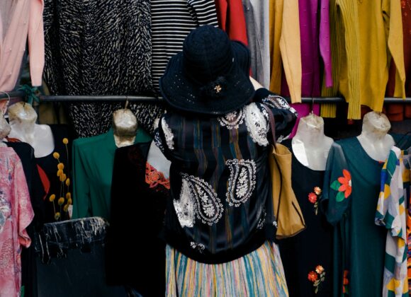 Why the Travel smith A Reversible Dress is a Must-Have A person wearing a black hat and colorful clothes browses sweaters hanging on a rack at an outdoor market. The scene is vibrant and lively.
