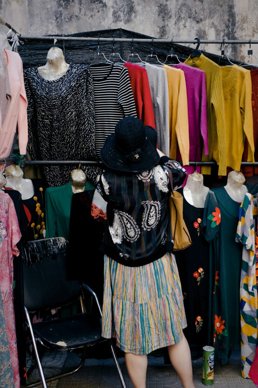 Why the Travel smith A Reversible Dress is a Must-Have A person wearing a black hat and colorful clothes browses sweaters hanging on a rack at an outdoor market. The scene is vibrant and lively.