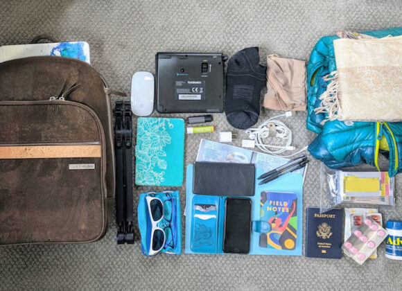 Packing for Europe in Fall: What to Wear and How to Travel Smart A neatly arranged travel essentials flat lay on carpet. Items include a brown backpack, blue and black clothing, tech gadgets, toiletries, sunglasses, documents, and medicine, conveying readiness and organization.
