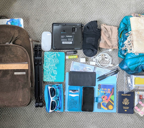 A neatly arranged travel essentials flat lay on carpet. Items include a brown backpack, blue and black clothing, tech gadgets, toiletries, sunglasses, documents, and medicine, conveying readiness and organization.