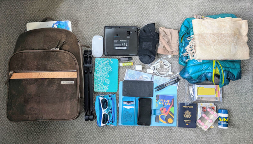 Packing for Europe in Fall: What to Wear and How to Travel Smart A neatly arranged travel essentials flat lay on carpet. Items include a brown backpack, blue and black clothing, tech gadgets, toiletries, sunglasses, documents, and medicine, conveying readiness and organization.