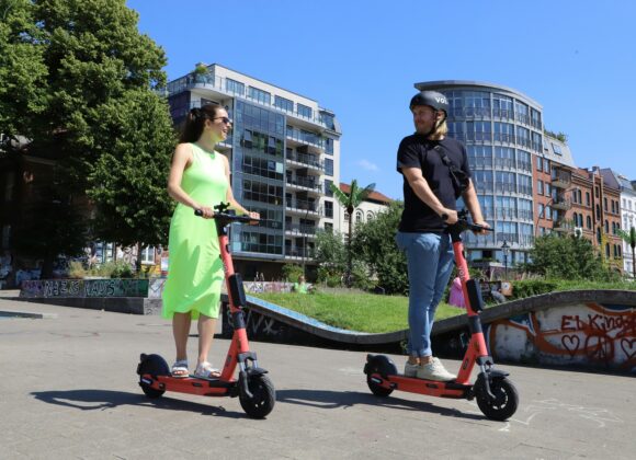 The Stylish Solution for Modern Mobility A woman and a man ride red e-scooters on a sunny day, smiling at each other. They're in an urban park with modern buildings and graffiti in the background.