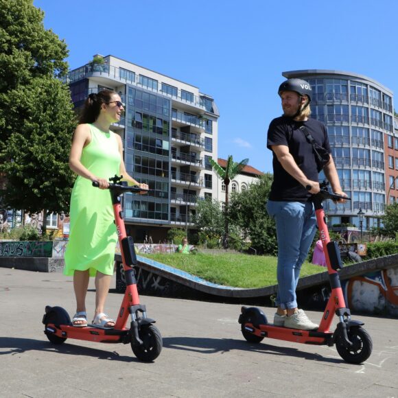 A woman and a man ride red e-scooters on a sunny day, smiling at each other. They're in an urban park with modern buildings and graffiti in the background.