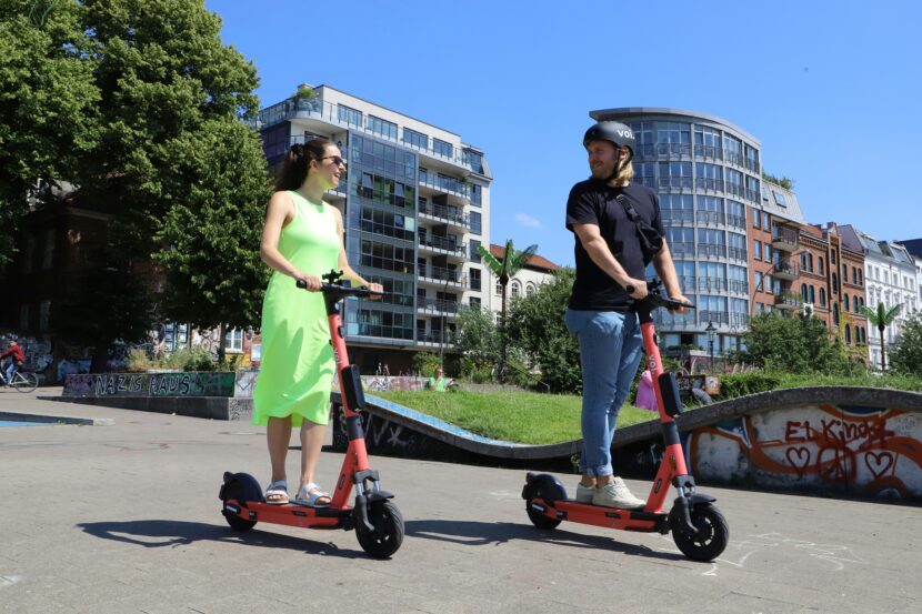 A woman and a man ride red e-scooters on a sunny day, smiling at each other. They're in an urban park with modern buildings and graffiti in the background.