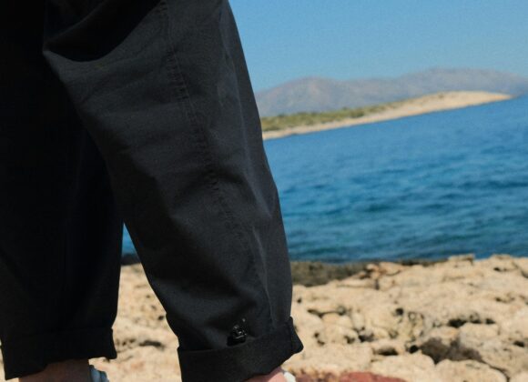 👟 Best Shoes for Greece: Your Ultimate Guide to Comfortable Walking Shoes for Greece 🇬🇷 Person in black pants and white sneakers with orange soles stands on a rocky shore. Background shows a calm blue sea and distant hills under a clear sky.