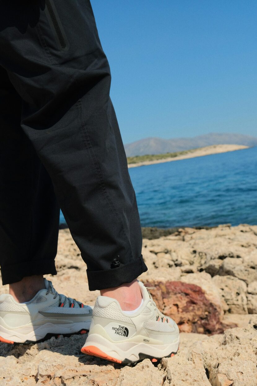 👟 Best Shoes for Greece: Your Ultimate Guide to Comfortable Walking Shoes for Greece 🇬🇷 Person in black pants and white sneakers with orange soles stands on a rocky shore. Background shows a calm blue sea and distant hills under a clear sky.