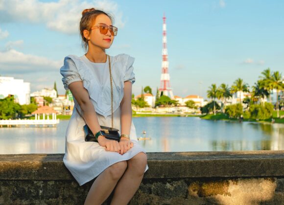 The Ultimate Guide to Fashion Travel Tumblers A woman in a white dress sits on a stone ledge by a tranquil lake, wearing sunglasses and flip-flops. The sky is blue with scattered clouds.
