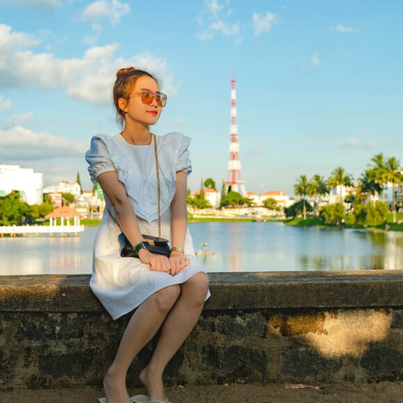 A woman in a white dress sits on a stone ledge by a tranquil lake, wearing sunglasses and flip-flops. The sky is blue with scattered clouds.