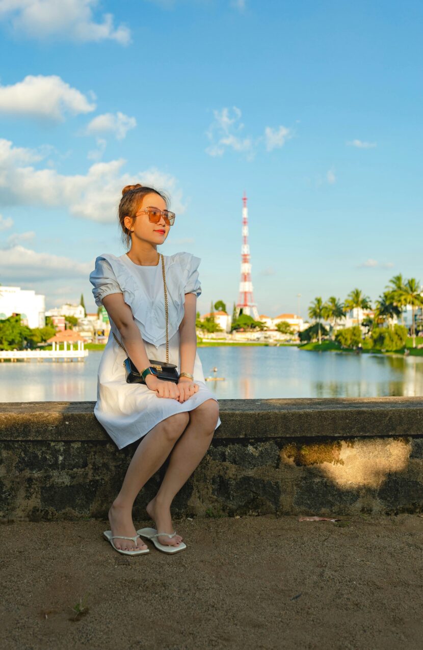A woman in a white dress sits on a stone ledge by a tranquil lake, wearing sunglasses and flip-flops. The sky is blue with scattered clouds.