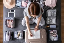 10-Step Guide to Packing for Different Climates Woman packing suitcase surrounded by travel essentials: clothes, shoes, hat, toiletries, tablet, and phone. She writes in a notebook, suggesting travel planning.