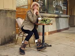 Women's Wide Width Boots: The Ultimate Style & Comfort Guide A person in winter clothes sits at a café table outdoors, smiling cheerfully. A coffee cup and flowers are on the table, with a "Café de la Paix" sign visible.