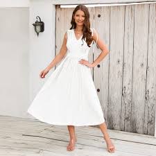 Women's White Dresses for Every Summer Escape A woman in a flowing white dress stands smiling by rustic wooden doors. The scene is bright and airy, conveying a sense of elegance and joy.