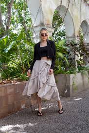 Women's Two-Piece Sets A woman in sunglasses stands outdoors, wearing a black jacket, crop top, and flowy skirt. She's surrounded by lush greenery and stone arches, exuding a relaxed, stylish vibe.