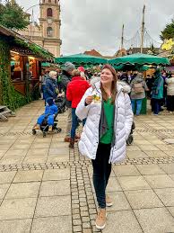 🎄 A Complete Guide + What to Wear to European Christmas Markets Woman in a white coat smiles and holds a drink in a bustling outdoor market. Green stalls and cobbled ground create a lively, festive atmosphere.