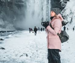 What to Pack for Iceland in March: Stay Warm, Dry, and Adventure A person in a pink coat and gray beanie holds a camera, standing on a snowy landscape near a waterfall. Others are in the background, creating a serene winter scene.