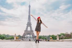 10-Step Packing Guide for a Trip to Paris and Italy Woman in a checkered dress and red beret joyfully poses with outstretched arms in front of the Eiffel Tower, under a partly cloudy sky in Paris.