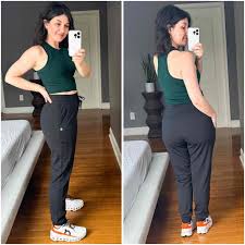 💪 Workout Leggings: Where Style Meets Strength A woman in a green sleeveless top and black pants poses in a bedroom. Wooden floor, white bed, and a gray wall create a relaxed, casual atmosphere.