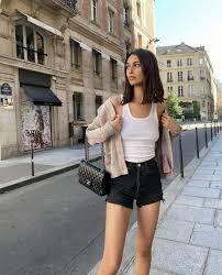 🇫🇷 Should You Wear Shorts in Paris—Ever Woman in casual attire stands on a city street, wearing a white tank top, black shorts, and a cardigan, with a purse. Urban setting, relaxed mood.