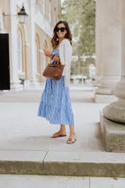 A woman in a blue dress and white cardigan stands on a stone path, holding a brown handbag. She wears sunglasses, exuding a casual, stylish vibe.