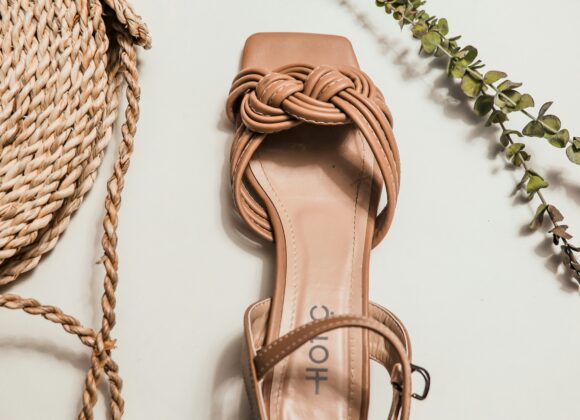 Naot Sabrina Sandals: Where Elegance Meets Everyday Comfort A beige sandal with a braided strap lies on a light surface, flanked by a coiled rope bag on the left and green sprigs on the right. Elegant and summery.