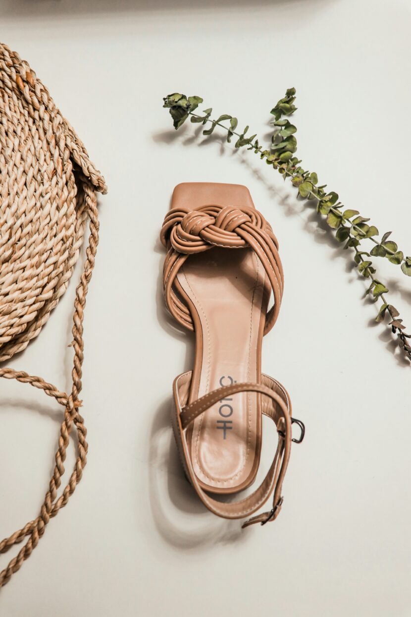 A beige sandal with a braided strap lies on a light surface, flanked by a coiled rope bag on the left and green sprigs on the right. Elegant and summery.
