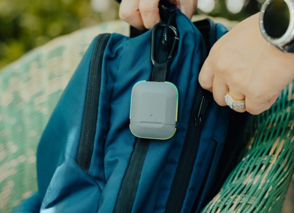 Why the Bags TLS Weekender Convertible Is a Game-Changer for Smart Travelers Hands attaching a small, square tracker to a blue backpack on a green wicker chair. The setting is outdoors, indicating travel or adventure.