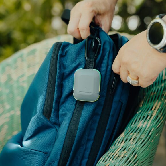 Hands attaching a small, square tracker to a blue backpack on a green wicker chair. The setting is outdoors, indicating travel or adventure.