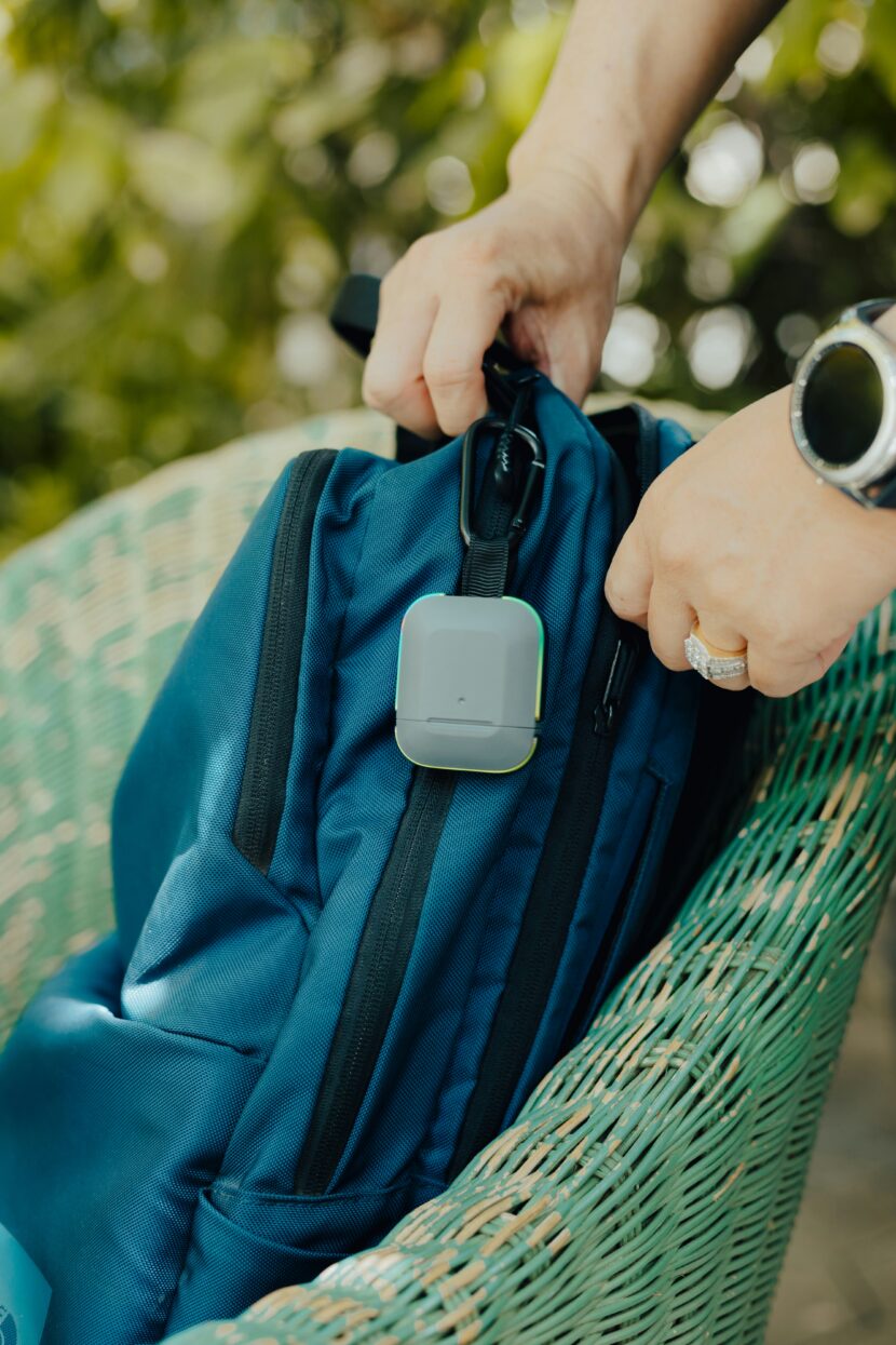Why the Bags TLS Weekender Convertible Is a Game-Changer for Smart Travelers Hands attaching a small, square tracker to a blue backpack on a green wicker chair. The setting is outdoors, indicating travel or adventure.