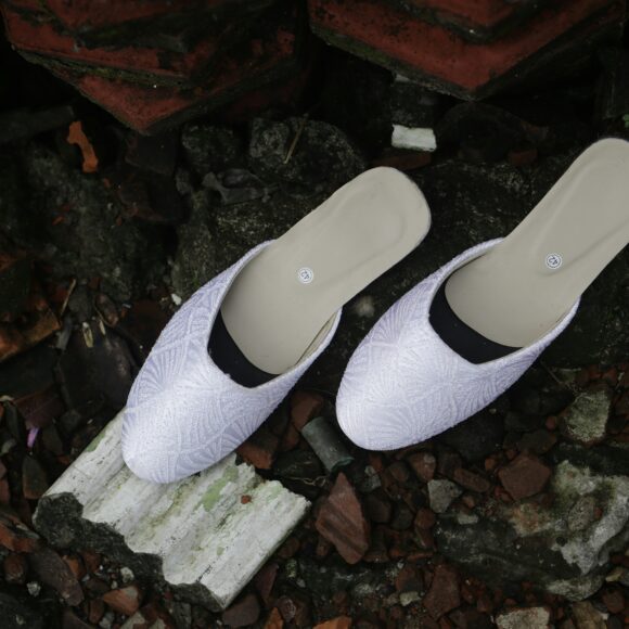 White slip-on shoes with a textured pattern sit on scattered, broken tiles and rocks. The scene conveys a contrast between elegance and decay.