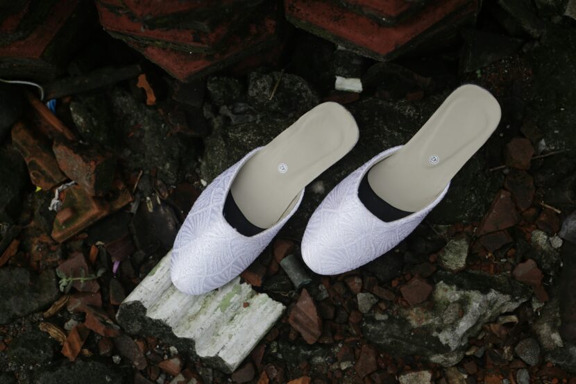 White slip-on shoes with a textured pattern sit on scattered, broken tiles and rocks. The scene conveys a contrast between elegance and decay.