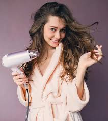 The Ultimate Guide to Dual Voltage Hair Dryers for Jet-Setting Style A woman in a fluffy robe smiles while holding a hairdryer. Her wavy hair flows freely, and the background is a soft purple, creating a cheerful ambiance.