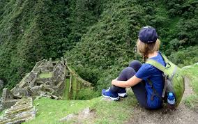Packing Guide for South America A person wearing a blue shirt and a green backpack sits on a hill overlooking ancient stone ruins surrounded by lush green forest, conveying a sense of adventure and exploration.