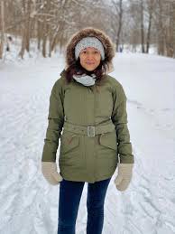A person stands on a snowy path in a forest, wearing a green parka with a fur-lined hood, gray headband, and gloves, conveying a serene winter mood.