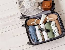 Packing for Cambodia: What to Wear and What to Leave Behind Open suitcase on white wooden floor containing neatly packed clothes, sandals, a wide-brimmed hat, sunglasses, and toiletries, suggesting a summer getaway.