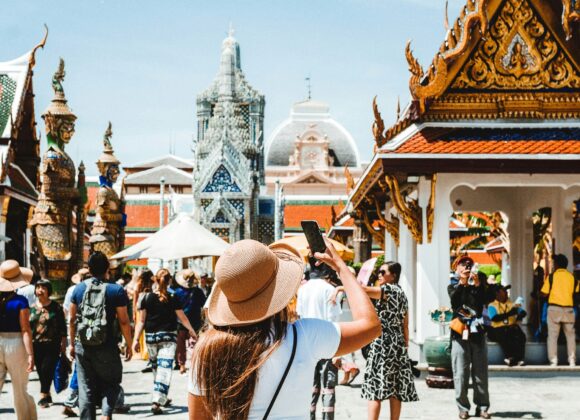 🧳 10-Step Packing Guide for Southeast Asia: Stylish, Smart & Culturally Aware A woman in a sun hat and blue pants takes a photo inside a busy temple courtyard in Thailand, surrounded by ornate architecture under a clear sky