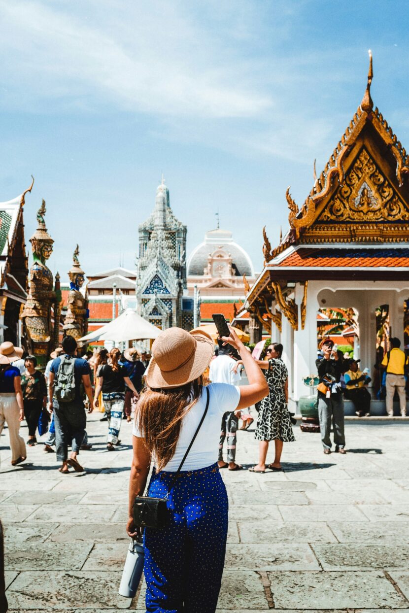 đź§ł 10-Step Packing Guide for Southeast Asia: Stylish, Smart & Culturally Aware A woman in a sun hat and blue pants takes a photo inside a busy temple courtyard in Thailand, surrounded by ornate architecture under a clear sky