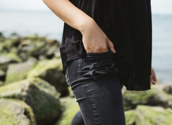 Most Comfortable Jeans for Women: The Ultimate Guide to Lightweight Black Jeans A person stands by a rocky, moss-covered shoreline, wearing a black top and ripped jeans, with one hand in their pocket. The mood is casual and relaxed.