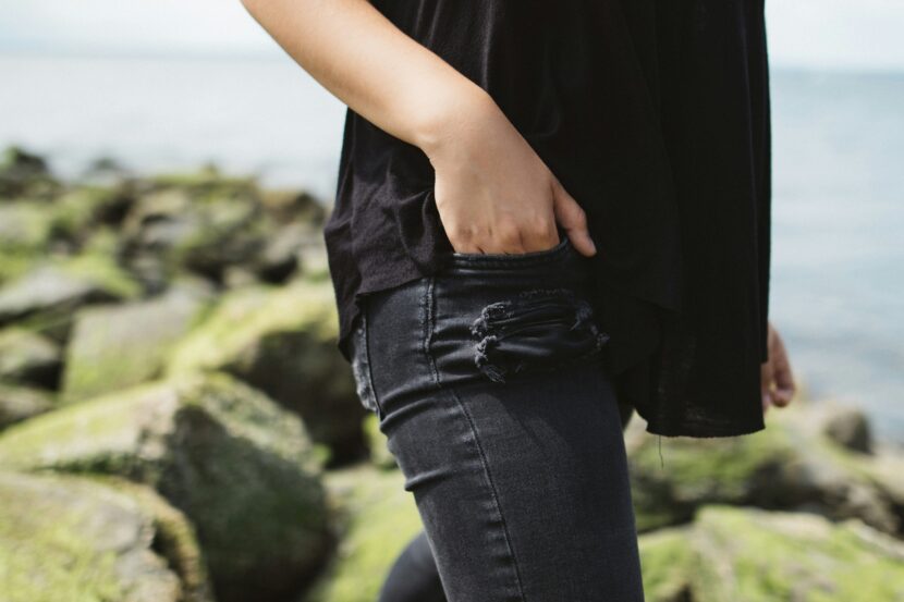 Most Comfortable Jeans for Women: The Ultimate Guide to Lightweight Black Jeans A person stands by a rocky, moss-covered shoreline, wearing a black top and ripped jeans, with one hand in their pocket. The mood is casual and relaxed.